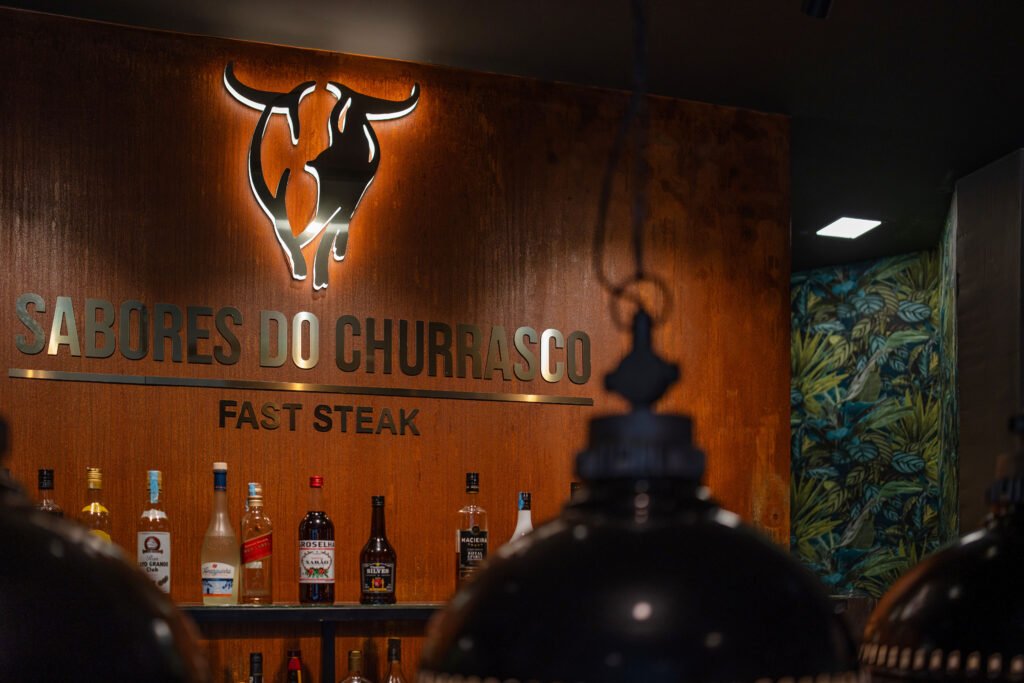 A decoração do restaurante na imagem é moderna e sofisticada, criando um ambiente acolhedor através de uma paleta de cores quentes, iluminação estratégica e uma mistura de materiais naturais e contemporâneos. O design interior é da autoria do The Creative & Co para a cadeia de restaurantes Sabores do Churrasco. A remodelação teve como objetivo criar um “espaço encantador” e elegante que sugere um local adequado tanto para refeições casuais quanto para ocasiões mais formais, complementando a oferta gastronómica de carnes e cozinha tradicional