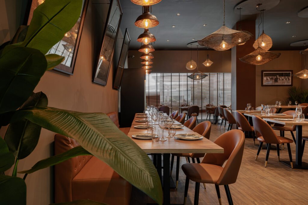 A decoração do restaurante na imagem é moderna e sofisticada, criando um ambiente acolhedor através de uma paleta de cores quentes, iluminação estratégica e uma mistura de materiais naturais e contemporâneos. O design interior é da autoria do The Creative & Co para a cadeia de restaurantes Sabores do Churrasco. A remodelação teve como objetivo criar um “espaço encantador” e elegante que sugere um local adequado tanto para refeições casuais quanto para ocasiões mais formais, complementando a oferta gastronómica de carnes e cozinha tradicional